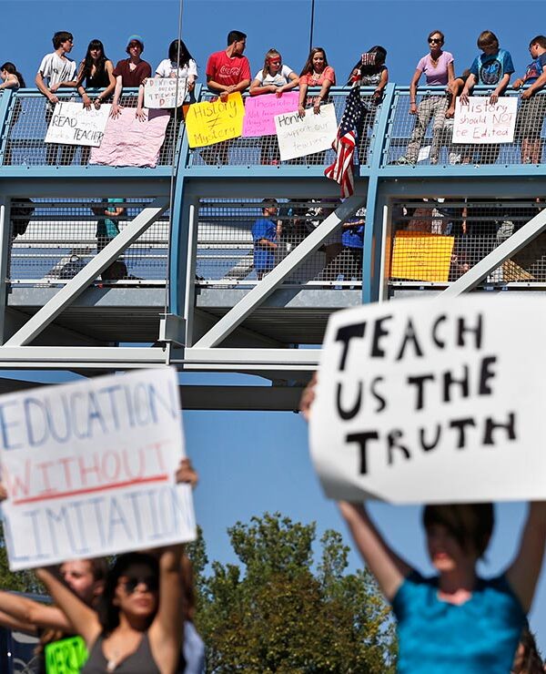 In the foreground (at an intersection,) two protesters carry signs with one reading "EDUCATION WITHOUT LIMITATION" and the other "TEACH US THE TRUTH", while in the background, other student demonstrators line an overpass protesting a proposal to...