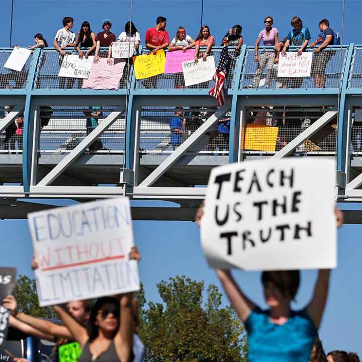 In the foreground (at an intersection,) two protesters carry signs with one reading "EDUCATION WITHOUT LIMITATION" and the other "TEACH US THE TRUTH", while in the background, other student demonstrators line an overpass protesting a proposal to...